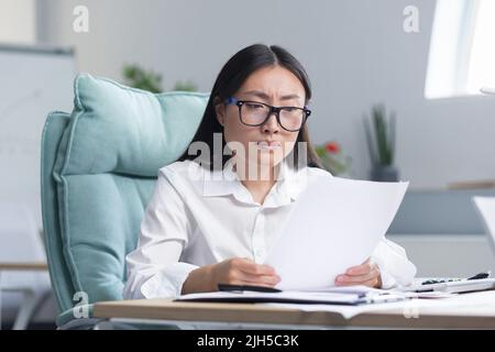 Die Fristen brennen.die junge, schöne asiatische Geschäftsfrau ist besorgt, sitzt im Büro am Schreibtisch, hält Dokumente in den Händen. Zeigt ihnen, hält ihren Kopf mit den Händen, weiß nicht, was zu tun ist. Stockfoto