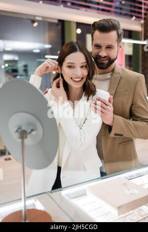 Ein fröhlicher Mann umarmte die Freundin mit einem Ohrring in der Nähe des Spiegels im Juweliergeschäft Stockfoto