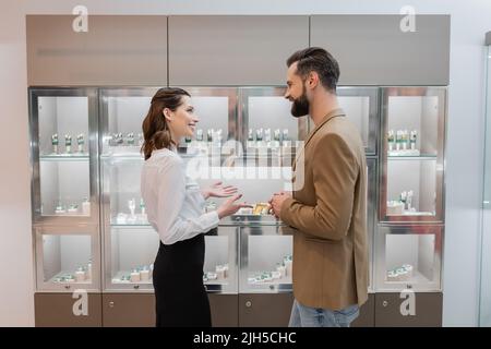 Seitenansicht eines lächelnden Verkäufers, der auf die Vitrine zeigt, während er mit dem Kunden im Juweliergeschäft spricht Stockfoto