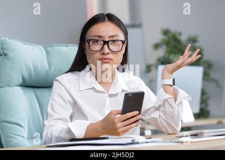 Besorgte junge, schöne asiatische Geschäftsfrau in Brille hält das Telefon in den Händen, schaut auf das Telefon, erhielt schlechte Nachrichten, breitet ihre Hände aus, überrascht, schockiert. Sitzen an einem Schreibtisch in einem modernen Büro. Stockfoto