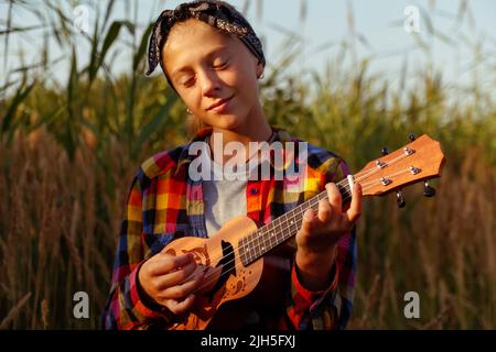 Defokussieren Mädchen mit Gitarre. Teen Mädchen zu Fuß auf Natur Hintergrund. Kleines Mädchen draußen auf grüner Wiese. Ukulelakkord. Sommerzeit. Menschen, Musiker. Stockfoto