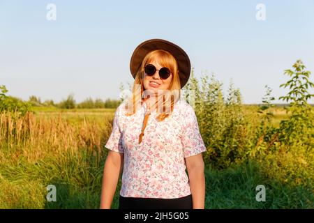 Unschärfe-Außenportrait einer lächelnden blonden Frau mittleren Alters in Hut und Sonnenbrille. Jugend-, Wellness- und Lifestyle-Konzept. 40s Frau auf Naturhinterg Stockfoto