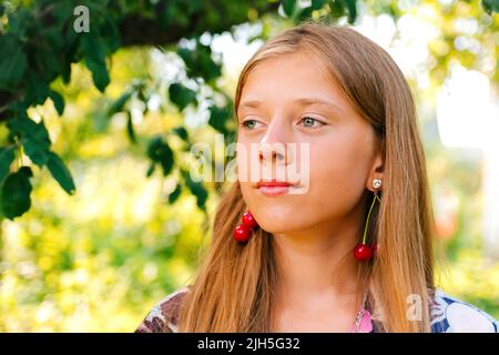 Defocus schöne blonde Teenager-Mädchen mit süßen Kirsche auf dem Ohr. Schönes lächelndes Teenager-Mädchen aus der Nähe, gegen das Grün des Sommerparks. Ohrringe für Kinder. Summ Stockfoto