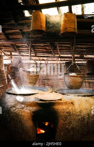 Die ländliche Produktion des Steinsalzes hing auf hölzernen Körben über kochendem Salzwasser in großen Gläsern. Sonnenlicht, das durch die Dachlöcher der Holzhütte scheint, die f berühren Stockfoto