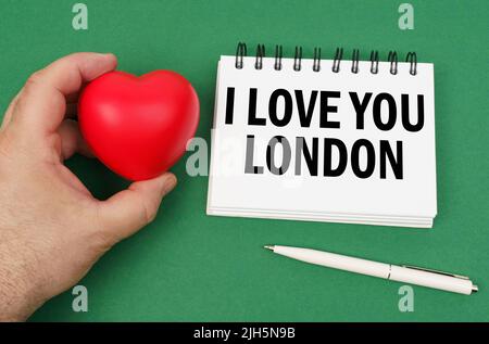 Auf der grünen Oberfläche befindet sich ein Stift, ein Notizblock mit der Aufschrift - I love you London. Ein Mann hat ein Herz in seinen Händen. Stockfoto