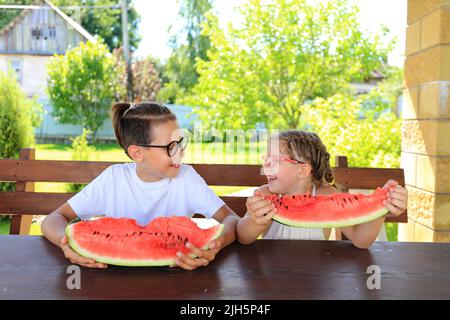 Kinder essen im Sommer große Wassermelone. Zufriedene Kinder mit einem großen Stück Wassermelone auf dem Hintergrund des Gartens Stockfoto