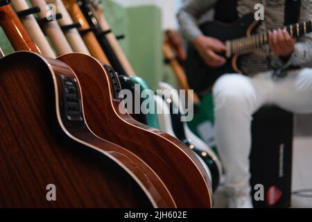 Viele elektrische und akustische Gitarren auf einem Ständer im Vordergrund mit einem Musiker im Hintergrund. Korpus der Gitarren. Gitarren im Music Store. Musiker spielt einen Sohn im Hintergrund mit schwarzer Gitarre. Stockfoto