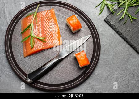 Lachsstücke, Küchenmesser und Rosmarinzweige auf Schneidebrett. Flach liegend. Brauner Hintergrund Stockfoto