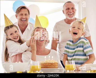 Kleines Mädchen, das die Augen der Mutter bedeckt, während es ihren Geburtstag feiert oder sie am Muttertag überrascht. Frau, die zu Hause mit ihrer Familie eine Party hat Stockfoto