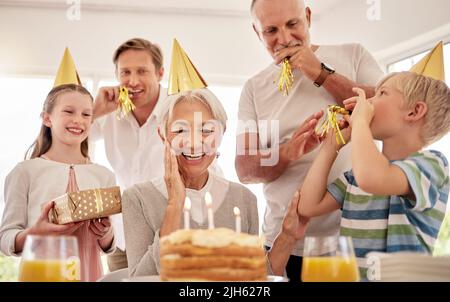 Ältere Frau, die ihren Geburtstag mit der Familie zu Hause feiert, trägt Partyhüte und pfeift. Oma schaut auf Geburtstagskuchen und schaut Stockfoto