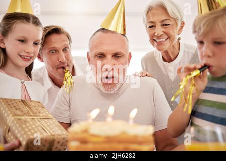 Ein älterer Mann, der mit seiner Familie zu Hause seinen Geburtstag feiert, trägt Partyhüte und pfeift. Großvater bläst Geburtstagskerzen aus und macht Stockfoto