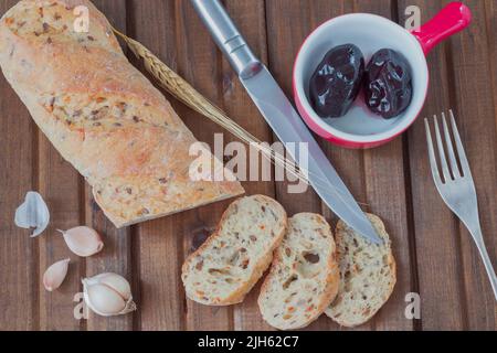 Weißbrot mit Sonnenblumenkernen in Stücke geschnitten. Eingelegte Pflaumen in einer Keramikschale. Messer. Gabel. Knoblauch und Zweig Gerste Stockfoto