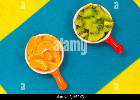 Scheiben von Mandarinen und Kiwi-Scheiben in Keramikplatten. Blau-gelber Hintergrund Stockfoto