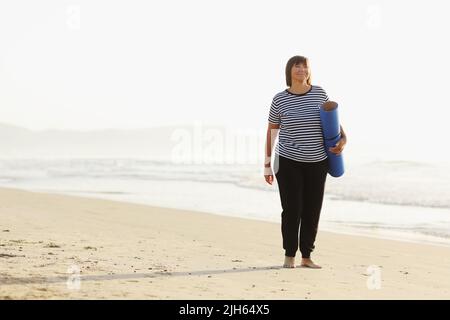 Frau im mittleren Alter, die eine Sportmatte hält und sich auf Yoga am Strand vorbereitet. Glückliche reife übergewichtige Frau, die am Meer trainiert Stockfoto