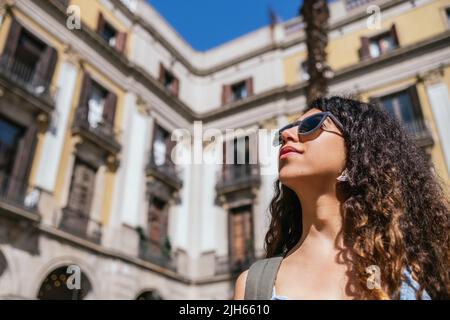 Junge, dunkelhaarige Frau mit Sonnenbrille schaut weg. Sie hat einen verschwommenen Hintergrund von alten Gebäuden. Stockfoto