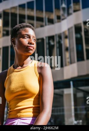 Vertikales Porträt einer jungen, stilvollen afrikanischen Frau mit kurzen Haaren und gelbem T-Shirt. Sie sieht ernst aus und schaut weg Stockfoto