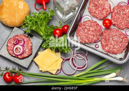 Grüne Zwiebeln, Käse, Wasserflasche. Brötchen mit Sesam, grünem Salat und Tomaten auf dem Tisch. Rohe Rindfleisch-Burger in der Pfanne. Flach liegend. Dunkler Hintergrund Stockfoto