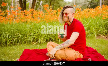 Junger Mann trinkt erfrischende Limonade und sitzt auf dem Teppich im Stadtpark. Entspannter Rüde mit Dreadlocks, der alkoholfreies Getränk durch Stroh trinkt, Durst löscht, Stockfoto