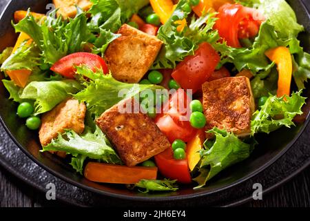 Nahaufnahme von gebratenem Tofu-Salat mit knackigen Salatblättern, grünen Erbsen, Tomaten, gelbem Paprika in schwarzer Schüssel Stockfoto