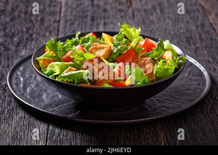 Gebratener Tofu-Salat mit knackigen Salatblättern, grünen Erbsen, Tomaten, gelbem Paprika in schwarzer Schüssel auf dunklem Holztisch Stockfoto