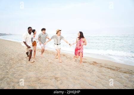 Fünf fröhliche Freunde, die Spaß am Strand haben, laufen im Sand zusammen, um einen glücklichen Wettbewerb zu führen. Junge Menschen genießen Sommerferien - Freundschaft Stockfoto