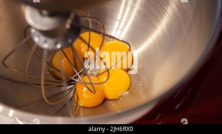 Rohe Eier in einer Metallschüssel mit stehendem Mixer. Eigelb mit Zucker in der Küchenarmatur zubereiten Stockfoto