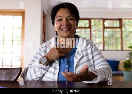Lächelnde, reife Frau mit biracial Haaren, die mit dem Finger auf den Hals zeigt, während sie am Tisch sitzt Stockfoto
