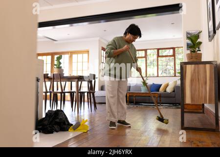 Volle Länge der reifen Biracial Frau mit kurzen Haaren fegenden Hartholzboden mit Besen zu Hause Stockfoto