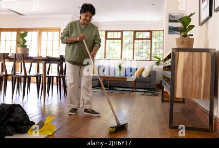 Volle Länge der Biracial reifen Frau mit kurzen Haaren fegenden Hartholzboden mit Besen zu Hause Stockfoto