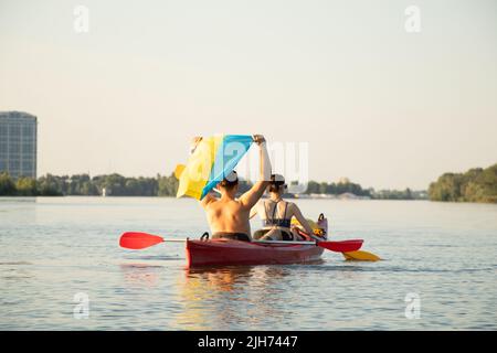 Ein Mädchen und ein Mann segeln in einem Kanu entlang des Dnjepr Flusses in der Stadt Dnepr in der Ukraine bei Sonnenuntergang mit der Flagge der Ukraine während des Krieges Stockfoto