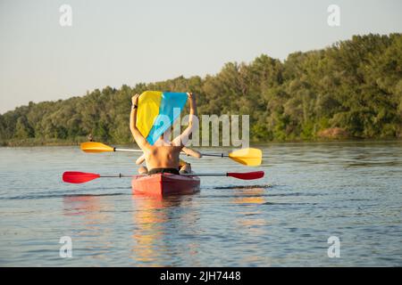 Ein Mädchen und ein Mann segeln in einem Kanu entlang des Dnjepr Flusses in der Stadt Dnepr in der Ukraine bei Sonnenuntergang mit der Flagge der Ukraine während des Krieges Stockfoto