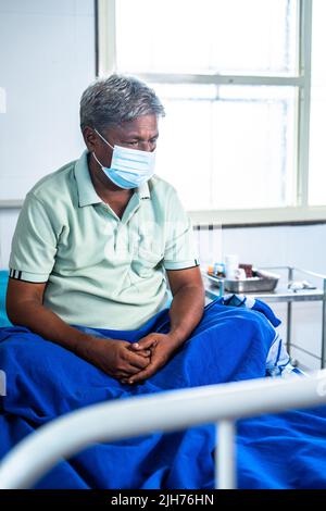 Vertikale Aufnahme eines gedankenvollen älteren Mannes mit medizinischer Gesichtsmaske, der auf einem Krankenhausbett sitzt - Konzept von Sorgen, Einsamkeit und Depression Stockfoto