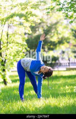 Eine 35-jährige Frau praktiziert Yoga in einem Stadtpark. Sie macht eine Yoga-Flexibilitätsübung. Vertikales Foto. Stockfoto