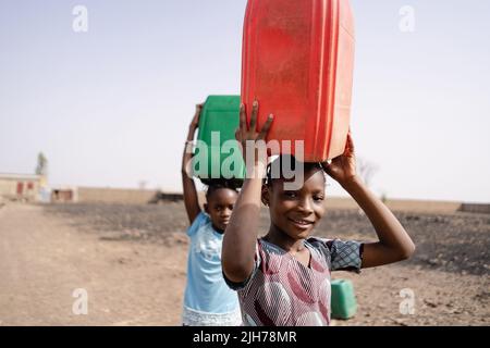 Nahaufnahme von zwei afrikanischen Mädchen, die Plastikwasserbehälter auf ihren Köpfen trugen und auf einer unbefestigten Straße in Richtung Dorf gingen Stockfoto