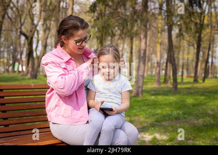 Auf einer Parkbank sitzend korrigiert die Mutter die Haare ihrer Tochter. Stockfoto