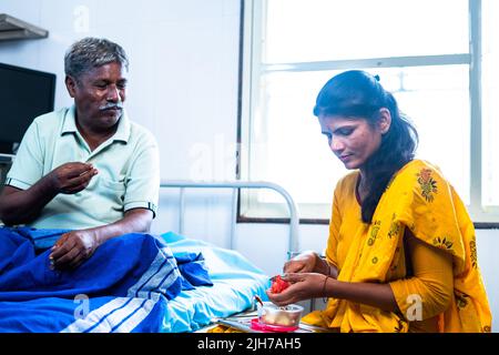 Glücklich lächelnde Tochter, die Früchte an wiedergeborgenen kranken Vater, während auf dem Bett im Krankenhaus - Konzept der Gesundheitsversorgung, Bindung und familiäre Unterstützung. Stockfoto