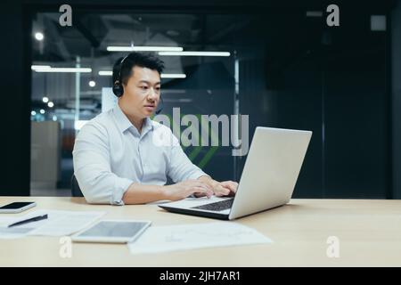 Junger gutaussehender asiatischer Geschäftsmann, der mit Kopfhörern und Mikrofon an einem Laptop arbeitet. Sitzen an einem Schreibtisch in einem modernen Büro. Ernst, konzentriert. Stockfoto