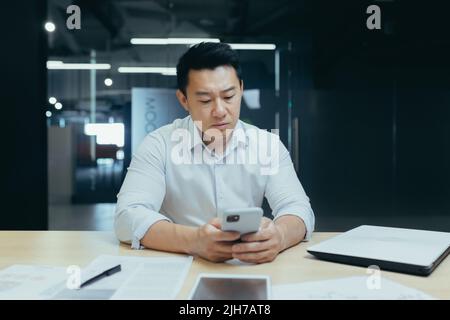 Schockiert und verärgert junger Mann asiatische Geschäftsmann Spiele auf Handy für Geld bei der Arbeit, verloren, erhielt schlechte Nachrichten durch Nachricht. Traurig am Schreibtisch in einem modernen Büro sitzen. Stockfoto