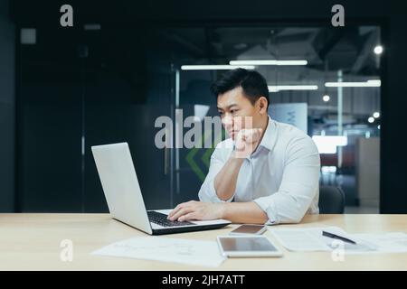 Ein junger, gutaussehender Mann, ein asiatischer Geschäftsmann, arbeitet mit einem Laptop und tippt. Sitzen an einem Schreibtisch in einem modernen Büro Stockfoto