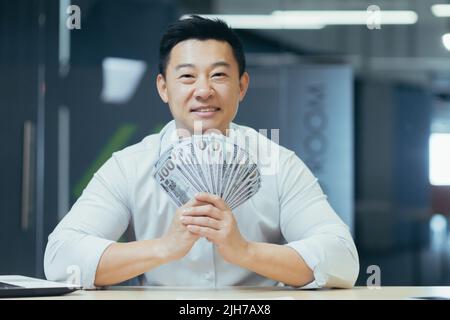 Ein glücklicher junger asiatischer Geschäftsmann, Freiberufler, Manager und Büroangestellter hält ein Bargeld in seinen Händen und winkt es. Er ist glücklich, zeigt auf die Kamera. Sitzen an einem Schreibtisch in einem modernen Büro Stockfoto