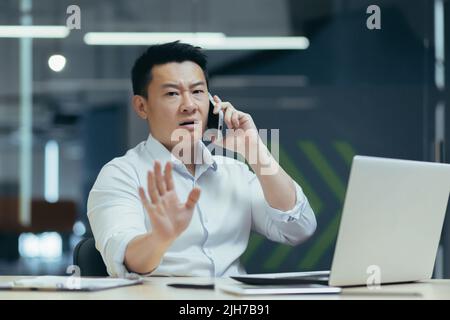 Besorgt und seriös junger männlicher asiatischer Geschäftsmann, der im Büro telefoniert. Sitzen an einem Tisch mit einem Laptop und Dokumenten. Es wird versucht, Probleme zu lösen Stockfoto