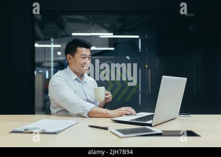 Kaffeepause. Ein junger Mann, ein asiatischer Geschäftsmann, ein Freiberufler, trinkt Kaffee aus einer Tasse, spricht von einem Laptop aus bei einem Videoanruf mit Familie und Freunden. Am Tisch in einem modernen Büro sitzen und lächeln. Stockfoto