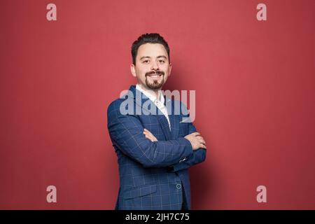Lächelnder bärtiger Geschäftsmann in blauem Anzug auf rotem Hintergrund Stockfoto