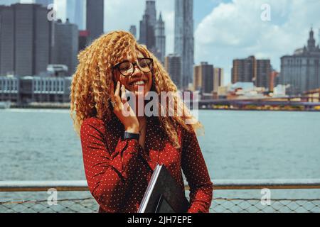 Porträt einer jungen erwachsenen Unternehmerin, tausendjährige Frau mit Brille und afrohaarigem Haar, die bei einem Telefonat im Freien lächelnd mit der Skyline von Manhattan, New York City, hinter dem Hudson River, spricht Stockfoto