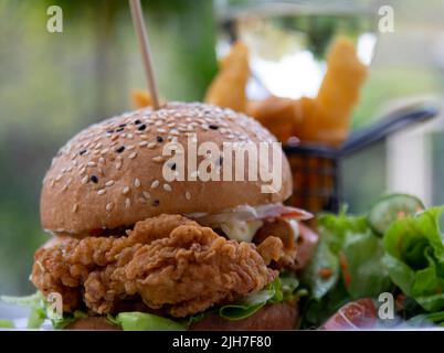 Ein knuspriger Chicken Burger mit einem Teller Salat und Getränken im Hintergrund Stockfoto