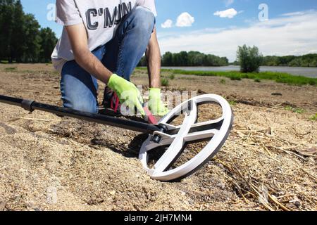 Ein Mann geht am Strand entlang und sucht nach einem Schatz mit einem drahtlosen Metalldetektor in der Hand und einem Pointer in der anderen Hand. Bäume im Hintergrund Stockfoto