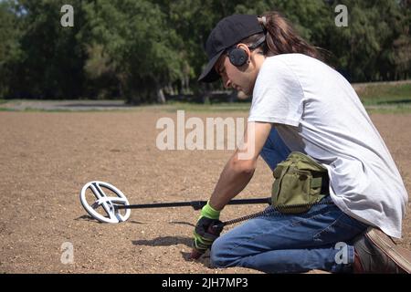 Ein Mann geht am Strand entlang und sucht nach einem Schatz mit einem drahtlosen Metalldetektor in der Hand und einem Pointer in der anderen Hand. Bäume im Hintergrund Stockfoto