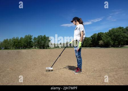 Ein Mann geht am Strand entlang und sucht nach einem Schatz mit einem drahtlosen Metalldetektor in der Hand und einem Pointer in der anderen Hand. Bäume im Hintergrund Stockfoto