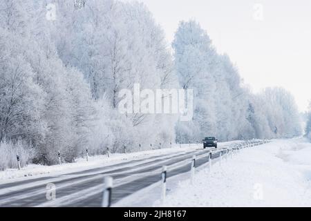 Ein dunkles Auto, das auf einer Autobahn fährt, umgeben von frostigen Bäumen und verschneiter Landschaft Stockfoto