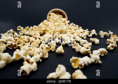 Popcorn wird aus einem braunen Papierbecher auf schwarzem Hintergrund in Nahaufnahme ausgestreut Stockfoto